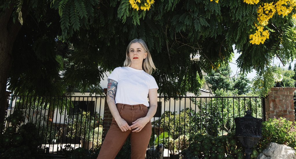 Portrait of the artist Debbie Bean standing under a tree with yellow flowers wearing a white t shirt and brown jeans with red lipstick on.