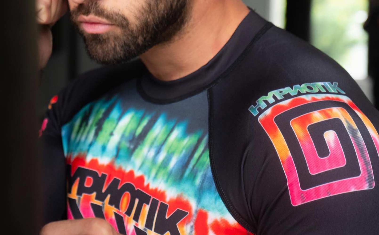 A closup of a man's shoulder, he's wearing a black performance shirt emblazoned with a tie-dyed Hypnotik logo.